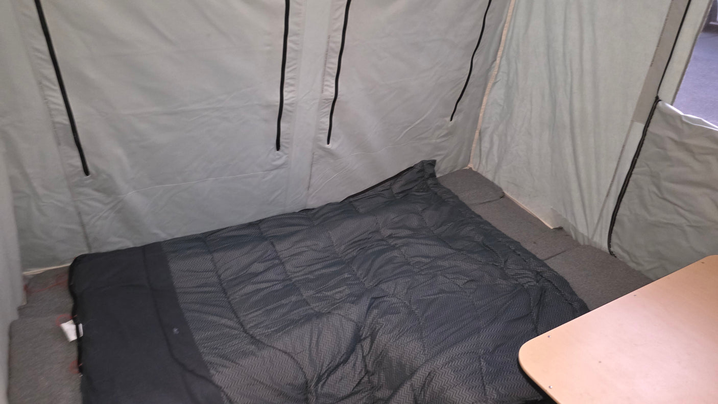 RV rental's compact interior: a dark sleeping bag on a gray bed, light fabric walls, and part of a wooden table.