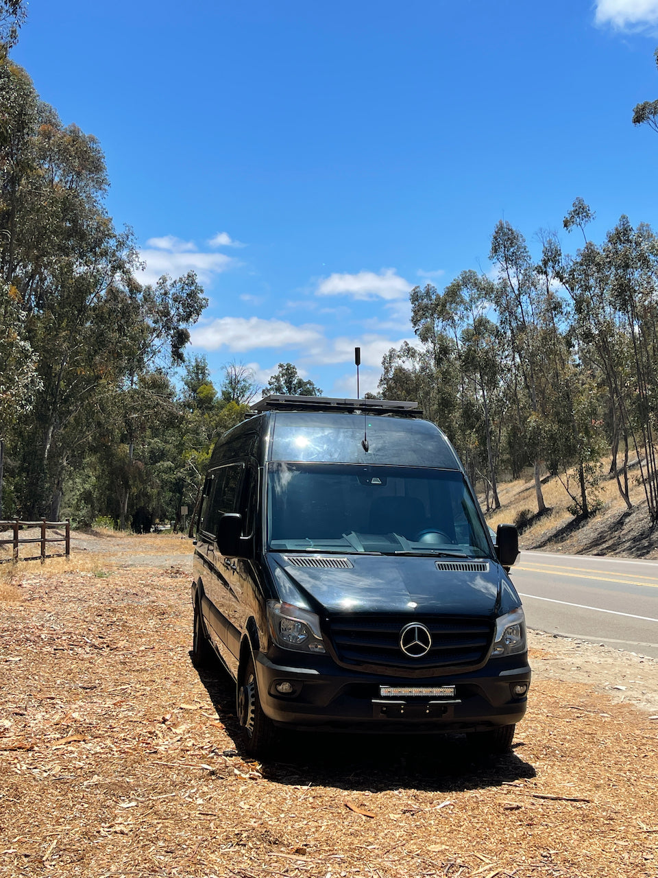 A black Mercedes-Benz Sprinter van, an ideal RV rental, parked on a sunny day next to a tree-lined road.