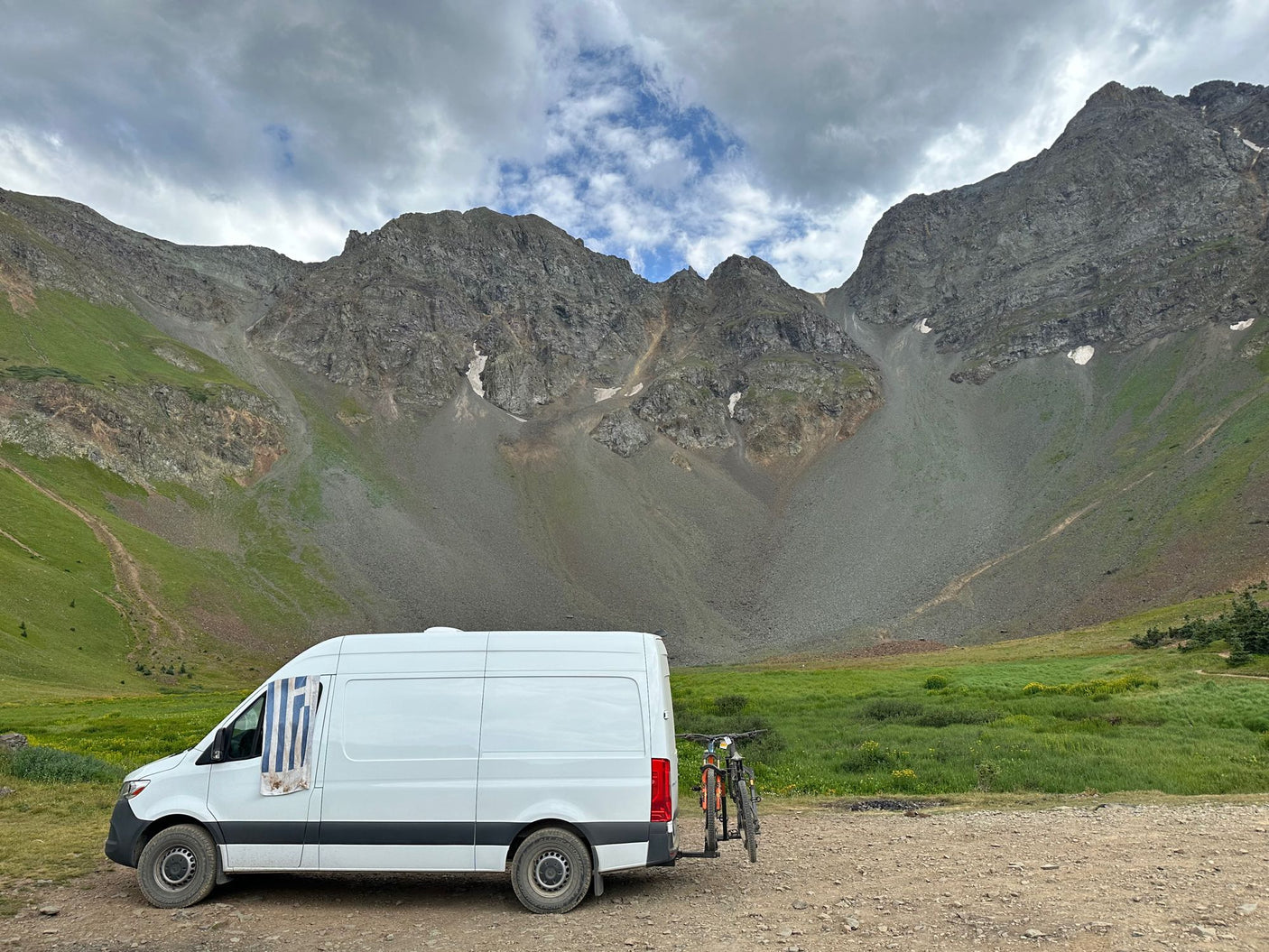 An RV rental camper van with bikes parked on a dirt road in a vast, rugged mountain landscape under cloudy skies.
