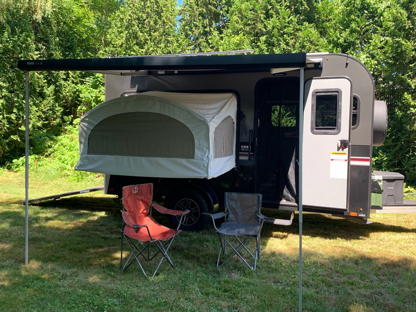 Grey rv rental camper with extended Thule awning, pop-out tent, and two chairs on grass amid green trees.