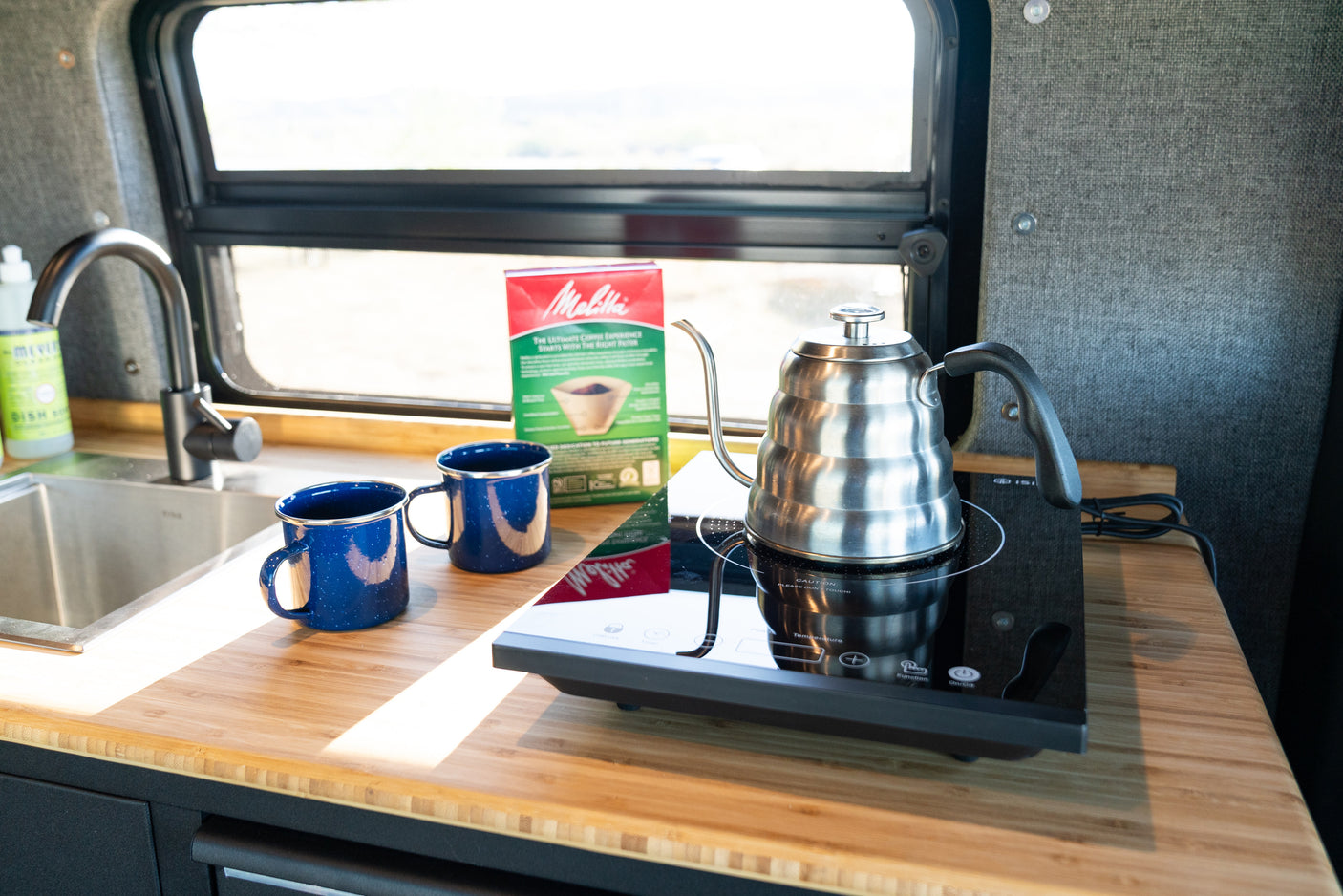 Enjoy morning coffee in this rv rental kitchen! Gooseneck kettle on induction cooktop, Melitta filters, sink, and blue mugs.