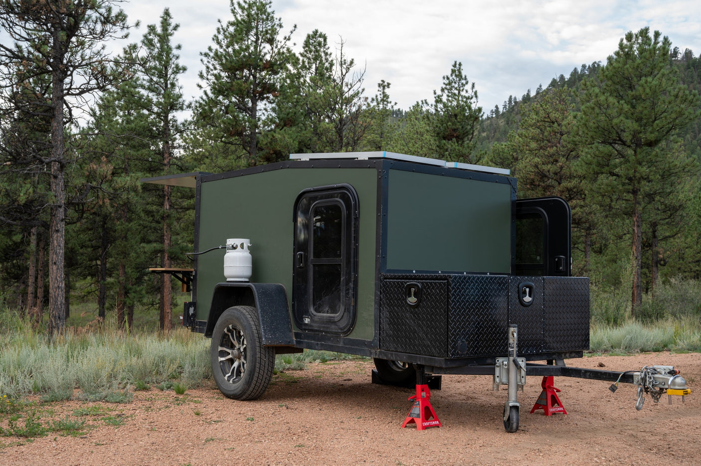 This rugged green camper is ready for your next **RV rental** adventure. Features propane, solar, and storage, set among pine trees.