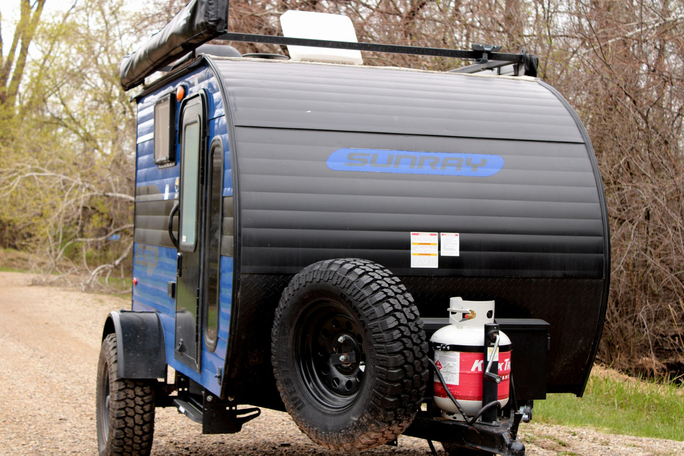 Blue Sunray teardrop rv rental with roof rack, ready for camping. Spare tire and propane tank visible on back.