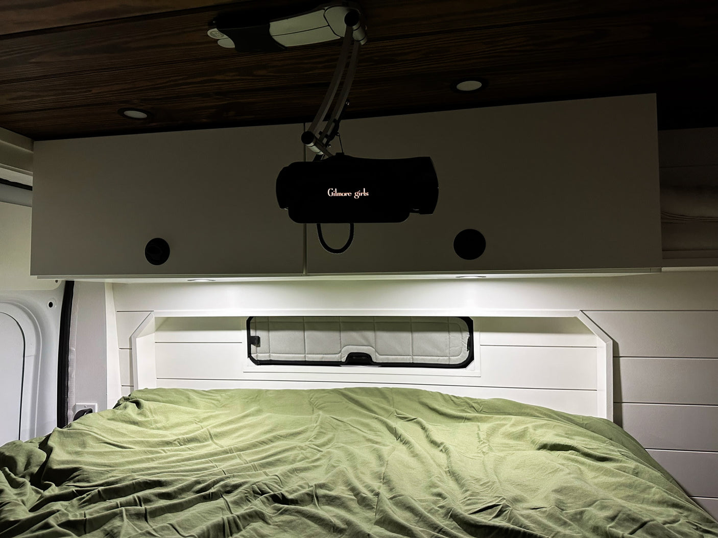Inside an RV rental, a bed with a green duvet and a projector displaying 'Gilmore Girls' on overhead cabinets for cozy viewing.