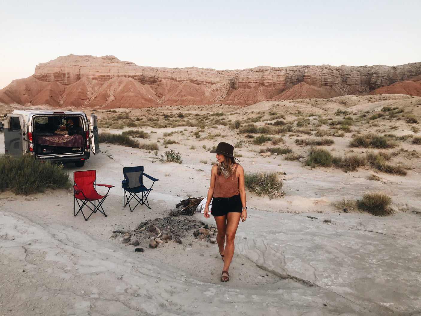 Woman walks by an RV rental camper van set up for desert camping with chairs and mountains in the background.