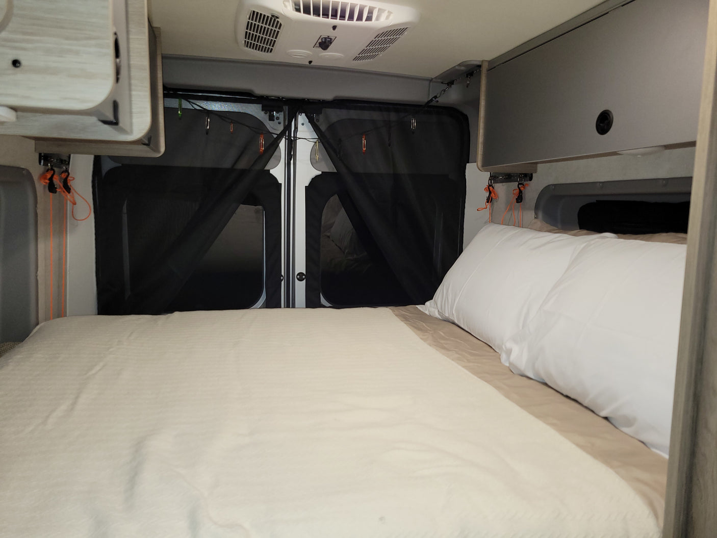 RV rental: Inside a camper van showing a cozy bed with white linens, black mesh screens on rear doors, overhead cabinets, and an AC unit.