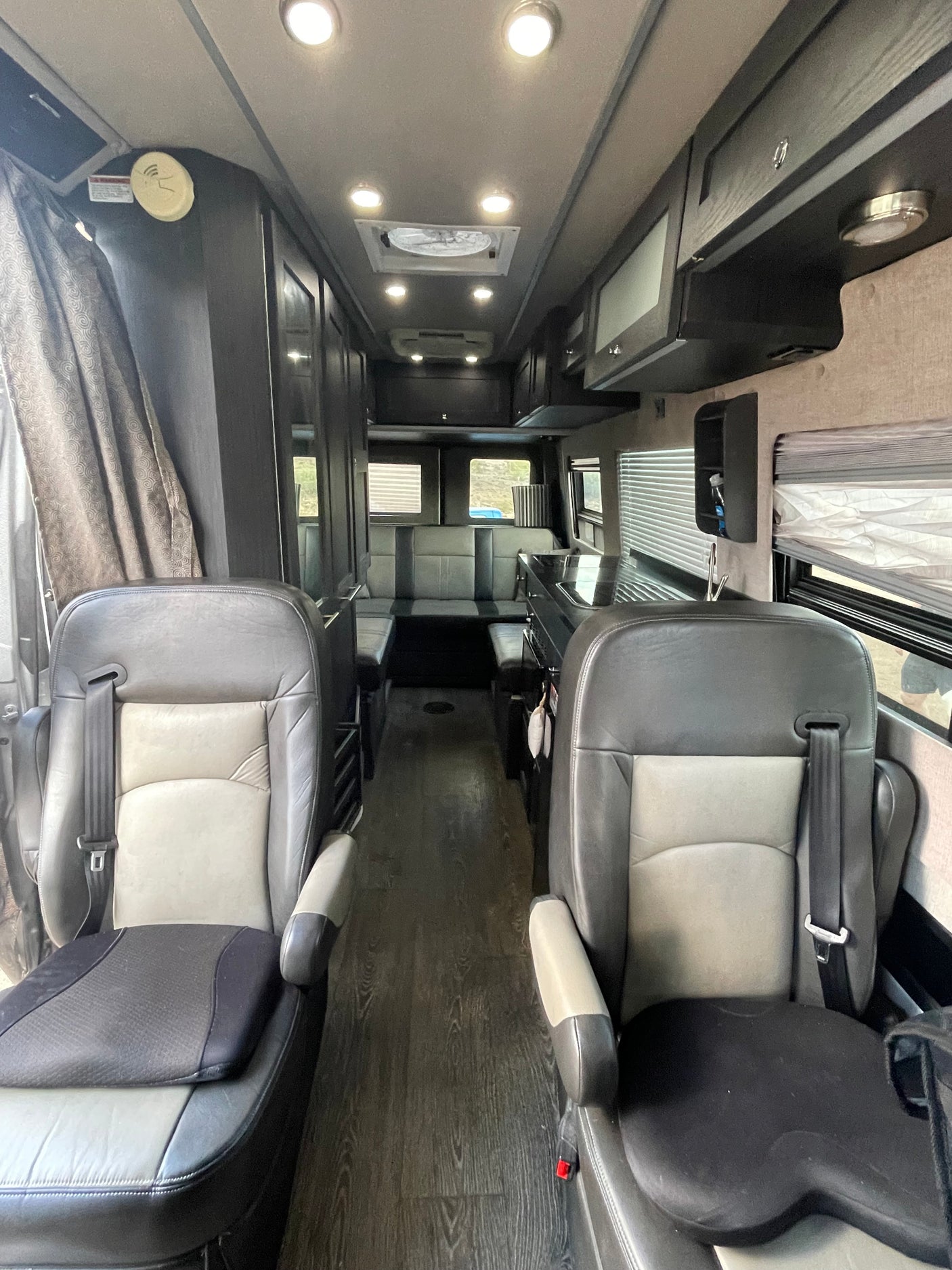 This RV rental interior features front captain's chairs, a kitchen, dinette, and dark wood-look flooring, with ample lighting.