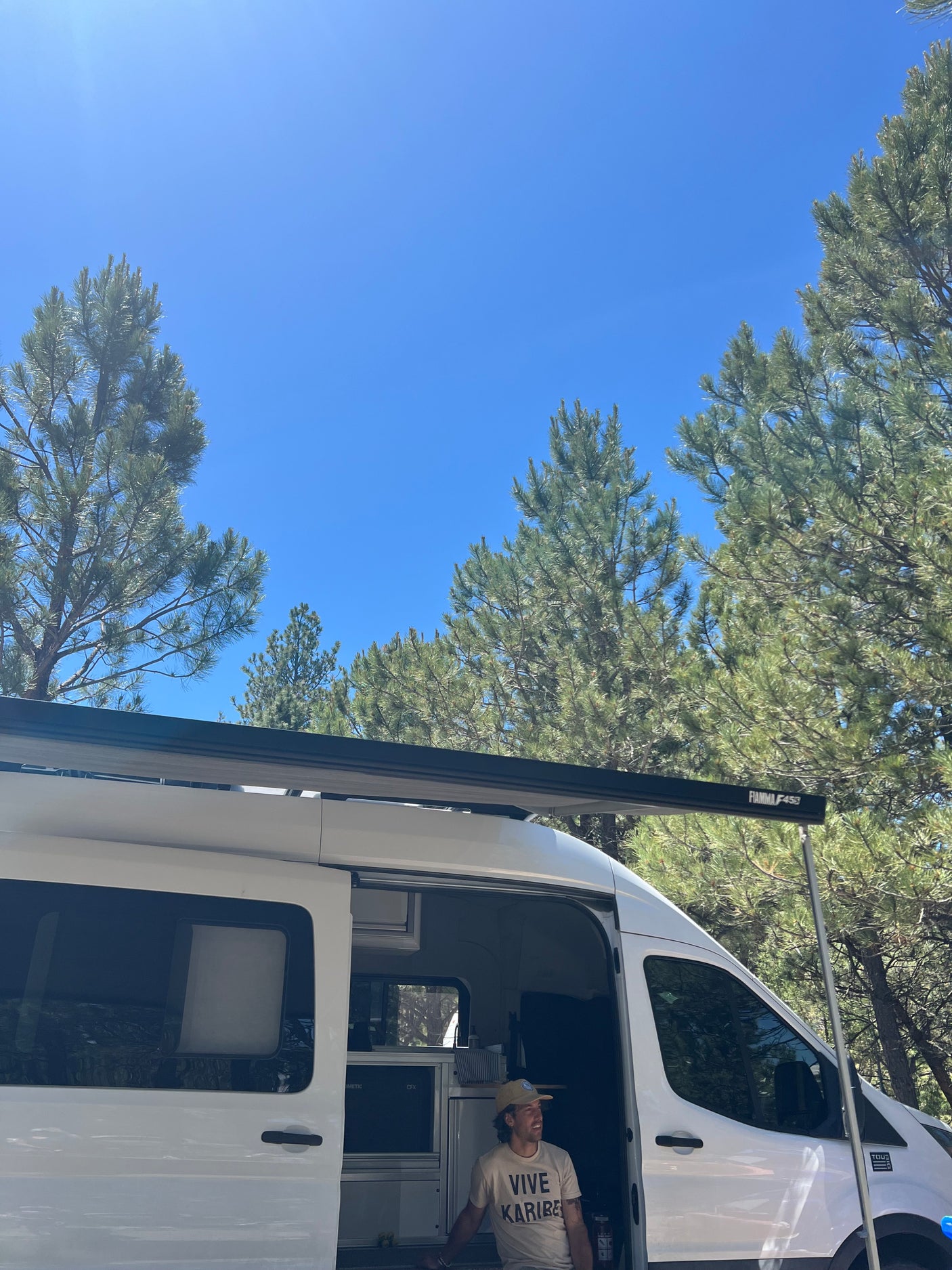 Man enjoying a sunny day from his RV rental camper van, parked amongst pine trees with its awning extended. Perfect for travel.