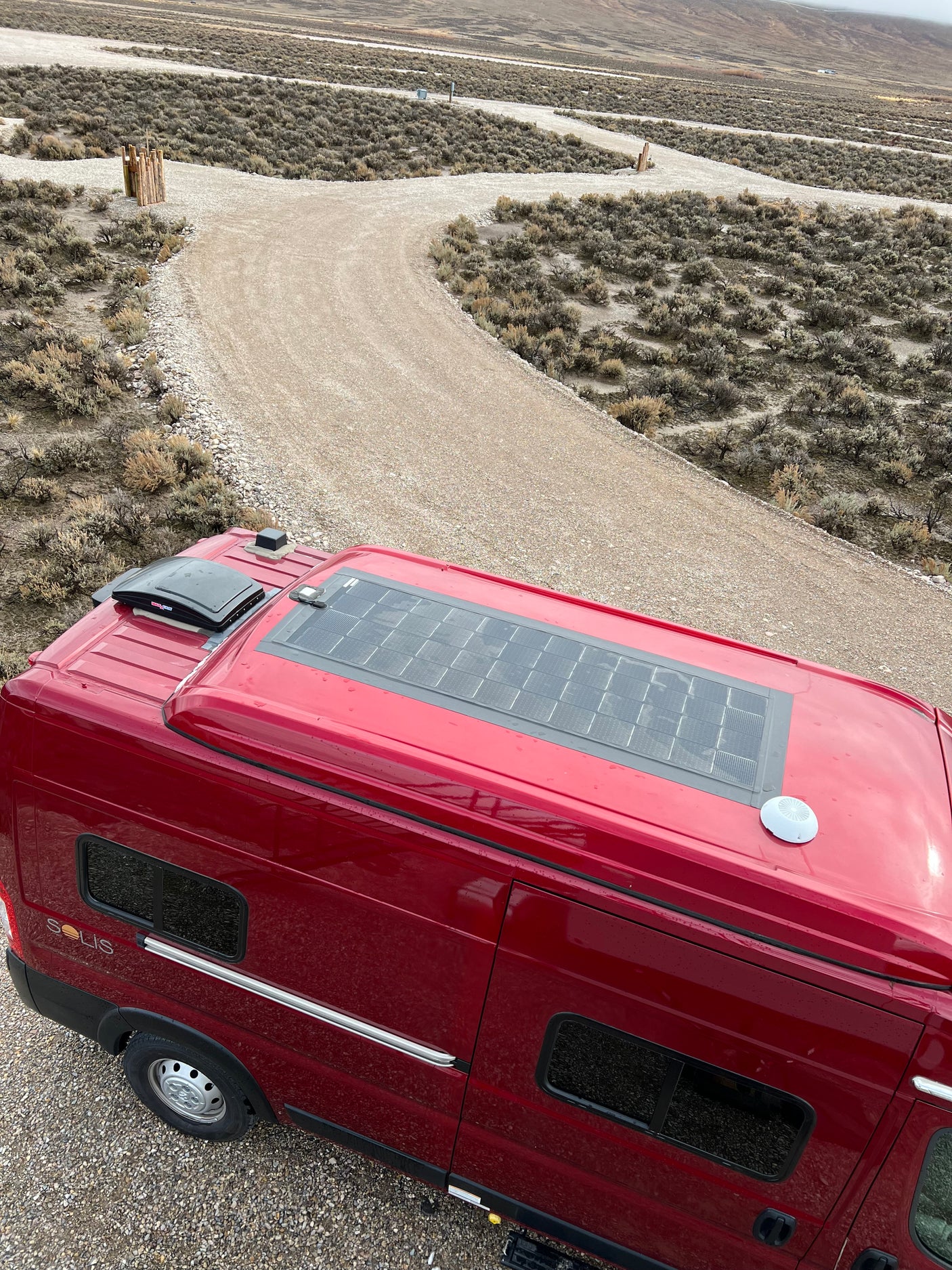 Overhead view of a red RV rental with solar panels on a remote gravel road, surrounded by dry, scrubby terrain.