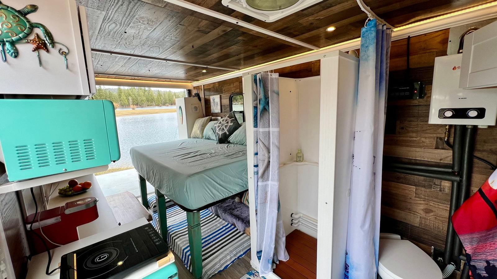This RV rental shows a cozy interior with a bed, kitchen, shower, and toilet, all with a beautiful lake view.