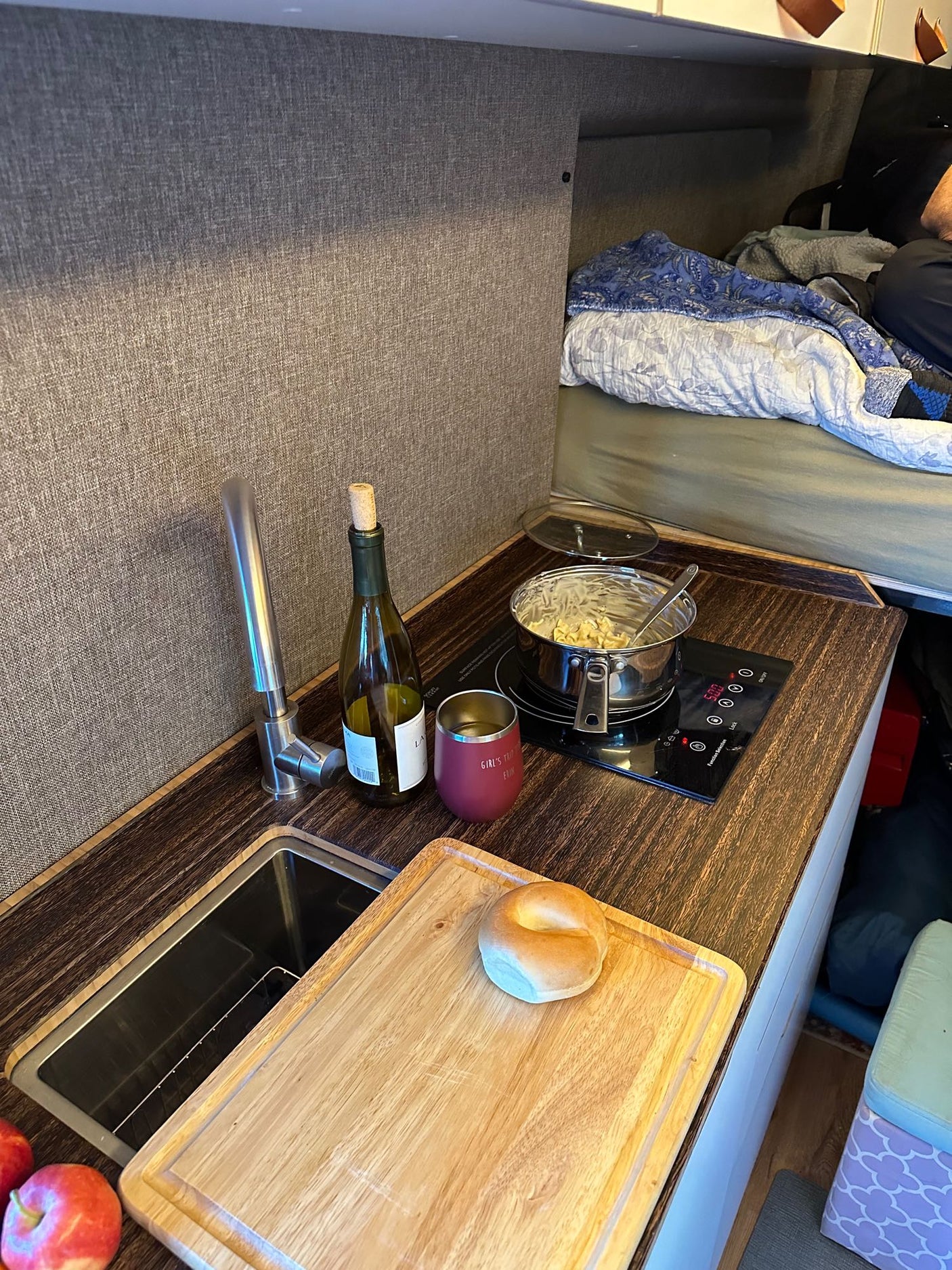 Cozy RV rental interior shows a compact kitchen with cooking pasta, sink, wine, and a bed in the background.