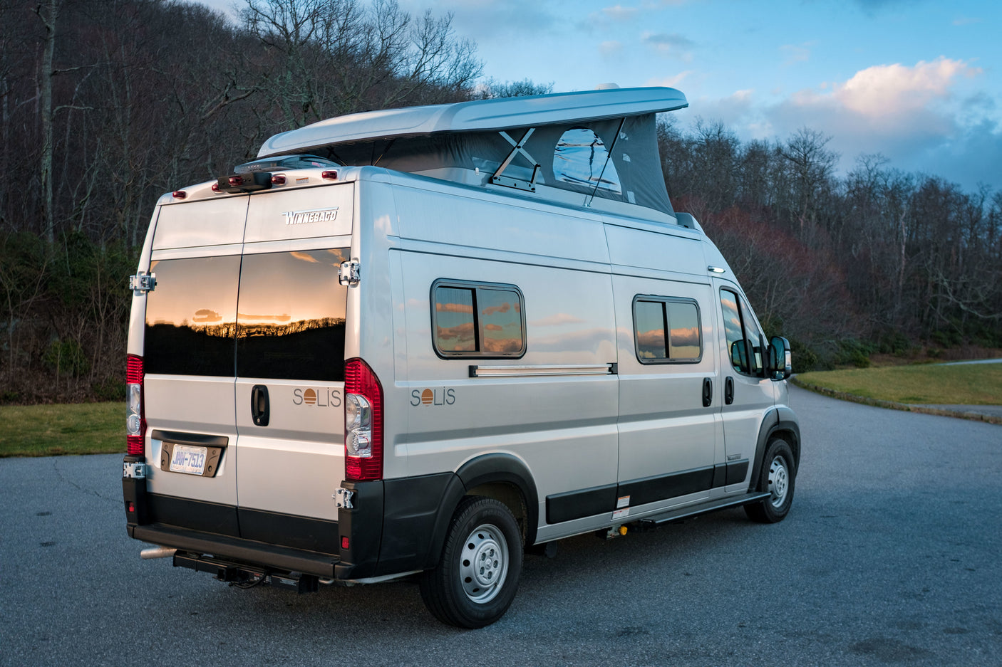Rear view of a silver Winnebago Solis pop-top camper van rv rental, parked on asphalt at dusk.