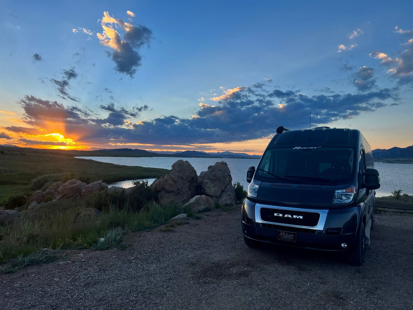 Experience a beautiful lake sunset with this RV rental. Black van parked on gravel, serene waters, vibrant sky.