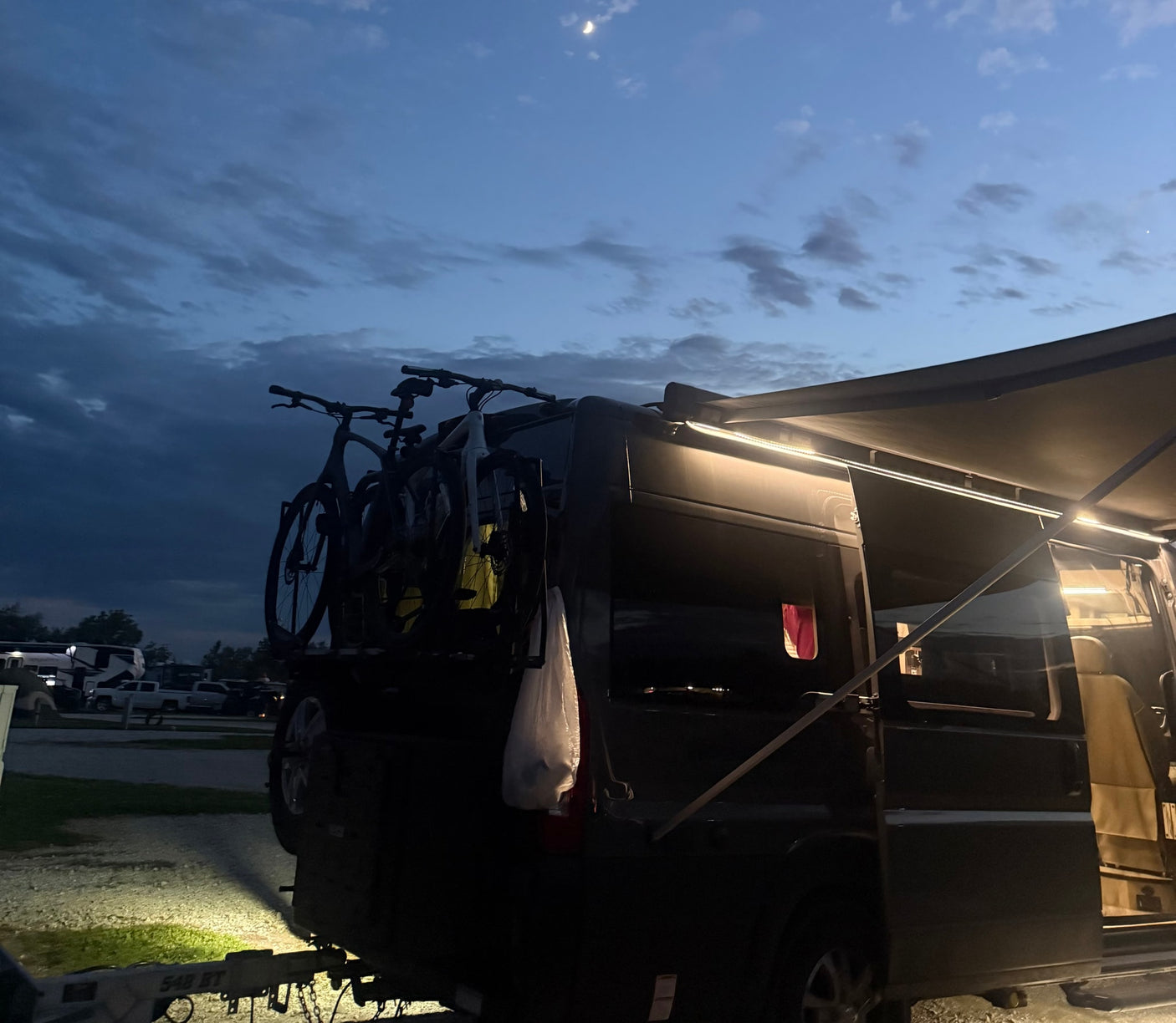 An RV rental at dusk, showcasing bikes on the rear, an illuminated awning, and a crescent moon. Ideal for outdoor adventures.
