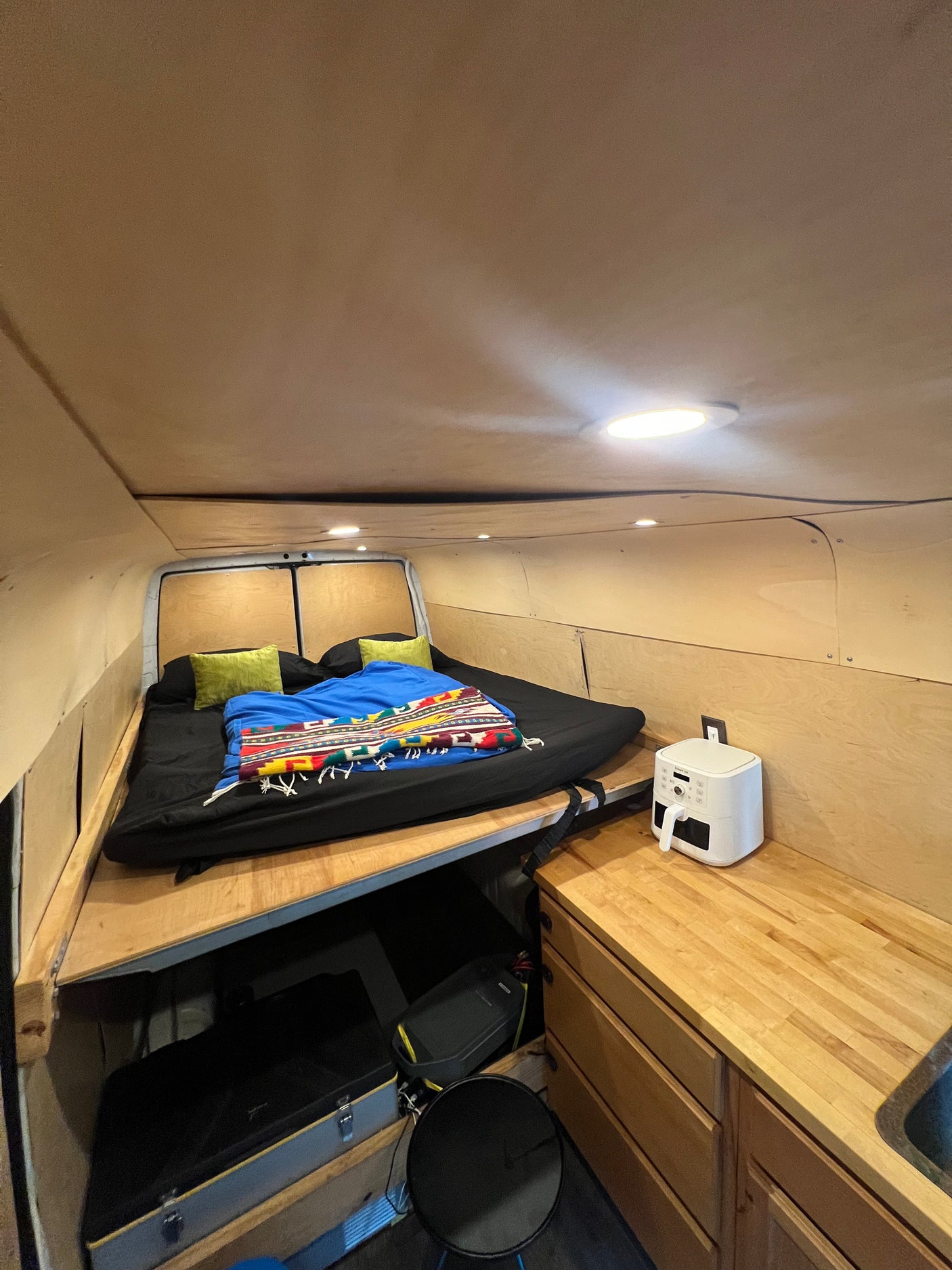 Inside this RV rental camper van: a bed with blankets, wooden walls/ceiling, recessed lights, and a kitchen counter with an air fryer.