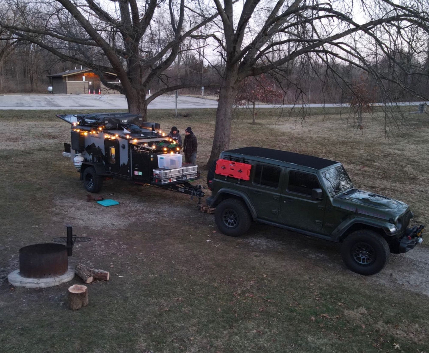 An overland Jeep tows a compact off-grid camper/rv rental, lit with string lights at a natural campsite with a fire pit.