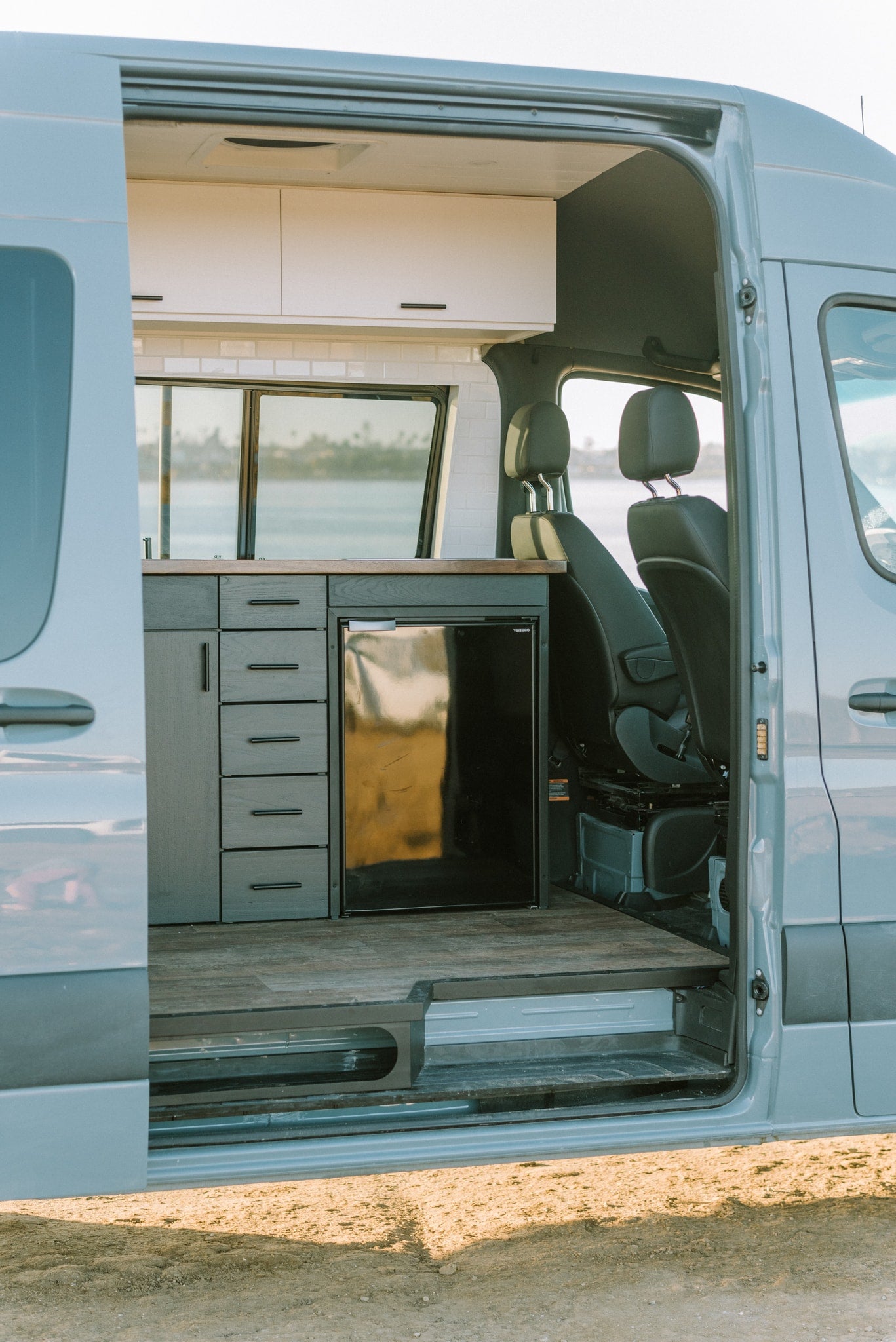 Modern rv rental interior: open side door reveals kitchen with dark cabinets, fridge, and wood floor, with water view. — photo 24