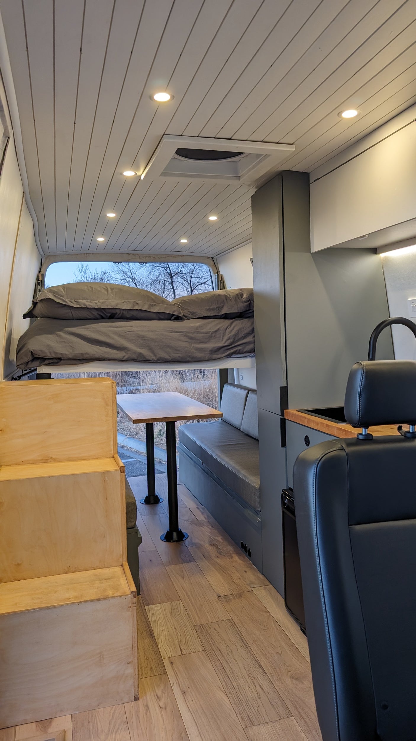 Inside a modern RV rental: A well-designed camper van interior with a raised bed, table, seating, and kitchen area. Wood floor & ceiling.