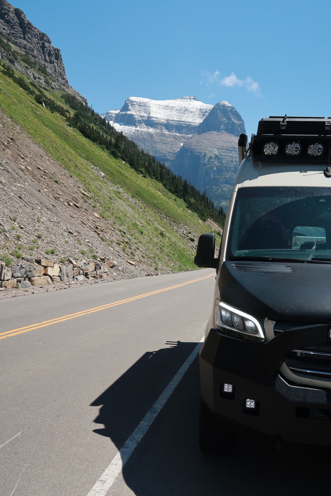 An RV rental is parked roadside, casting a long shadow, amidst green mountain slopes with distant snow-capped peaks under a blue sky.