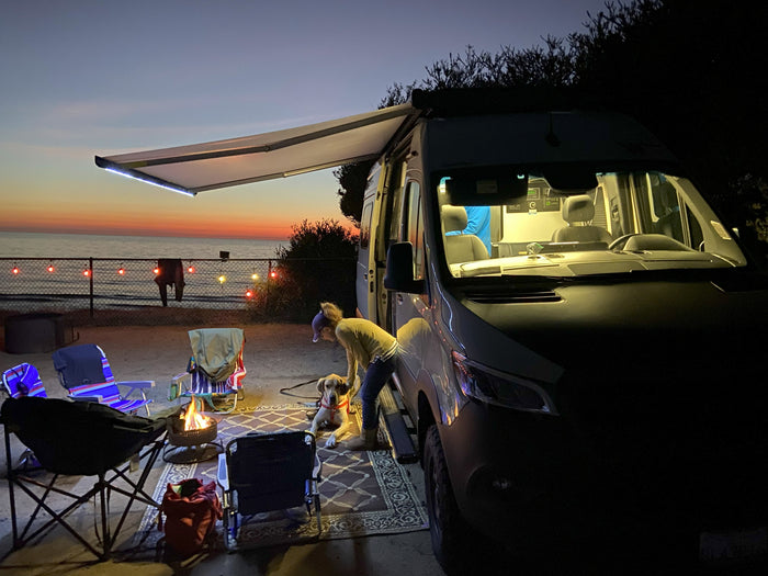 An RV rental campervan lights up a beach evening with a person, dog, and warm campfire at sunset.
