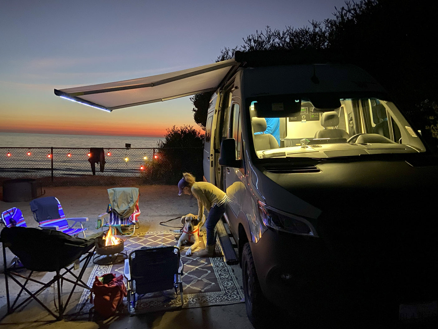 An RV rental campervan lights up a beach evening with a person, dog, and warm campfire at sunset.