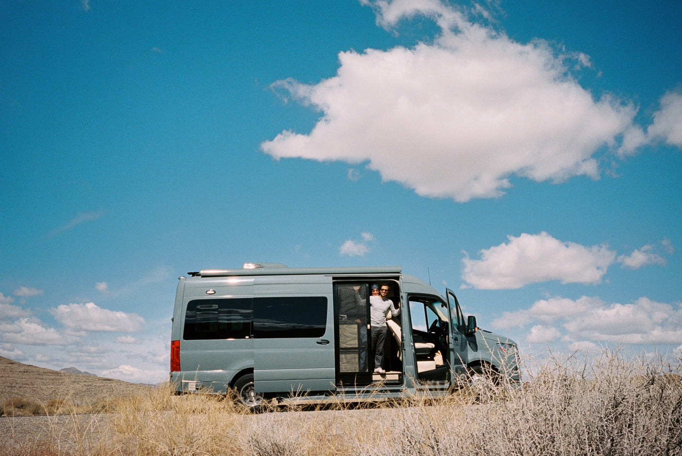 A man smiles from an RV rental van, parked in a sunny desert landscape with dry brush under a bright blue sky.