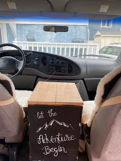 Interior view of an RV rental van, looking forward. Features front seats, dashboard, and a decorative "Let the Adventure Begin" sign.