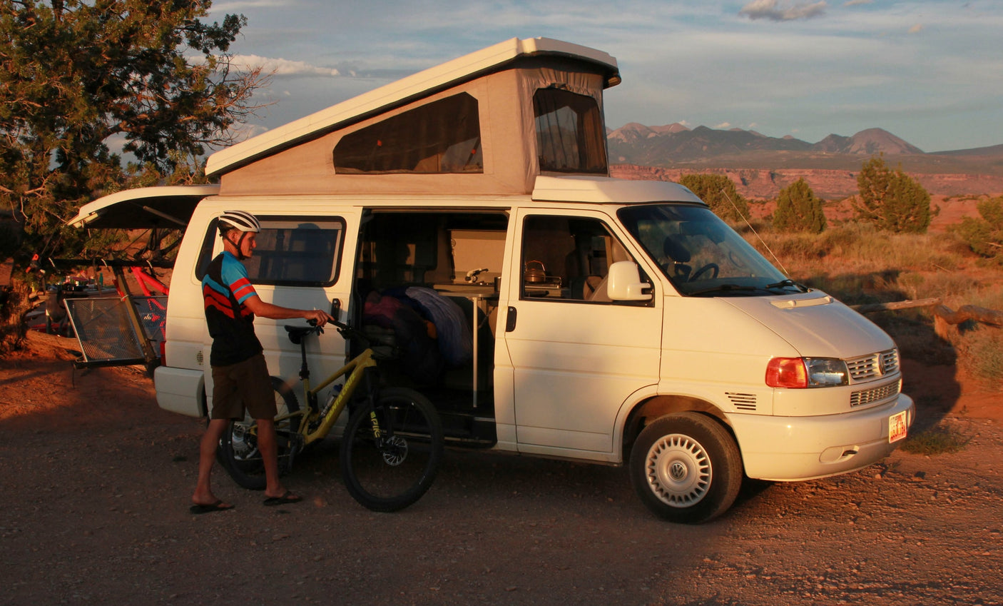Man with mountain bike by a white pop-top camper van in a desert sunset. An ideal RV rental for outdoor adventures.