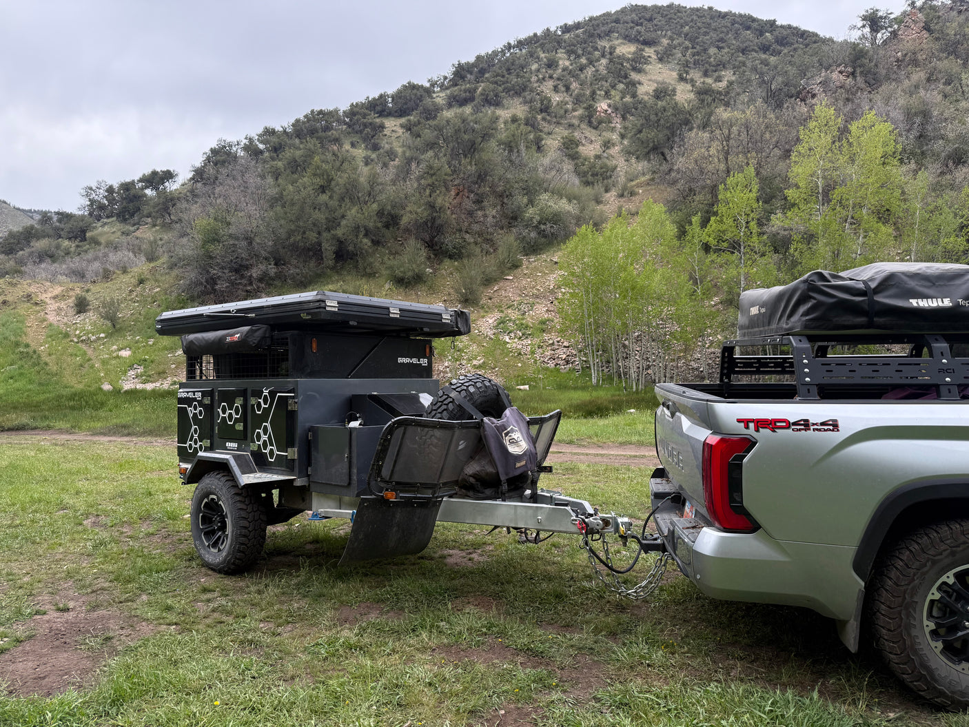 An overland RV rental trailer with rooftop tent hitched to a truck, parked in a grassy mountain area.