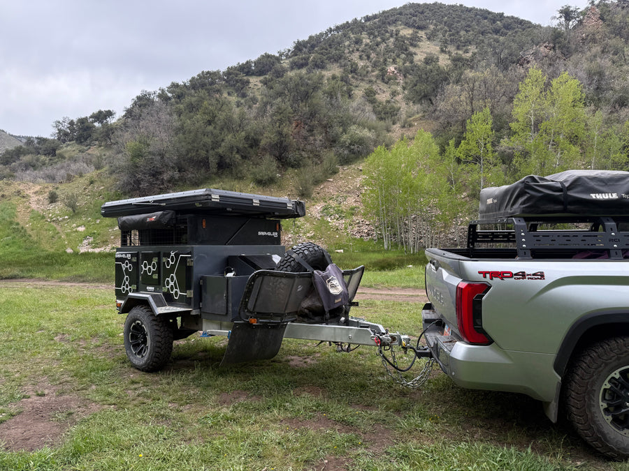 An overland RV rental trailer with rooftop tent hitched to a truck, parked in a grassy mountain area.