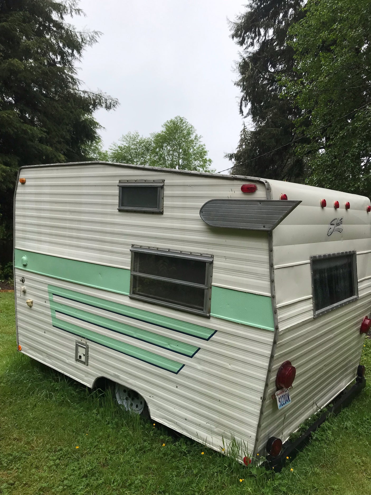 A vintage white & green travel trailer RV, ready for an rv rental, parked in a grassy yard under a cloudy sky.