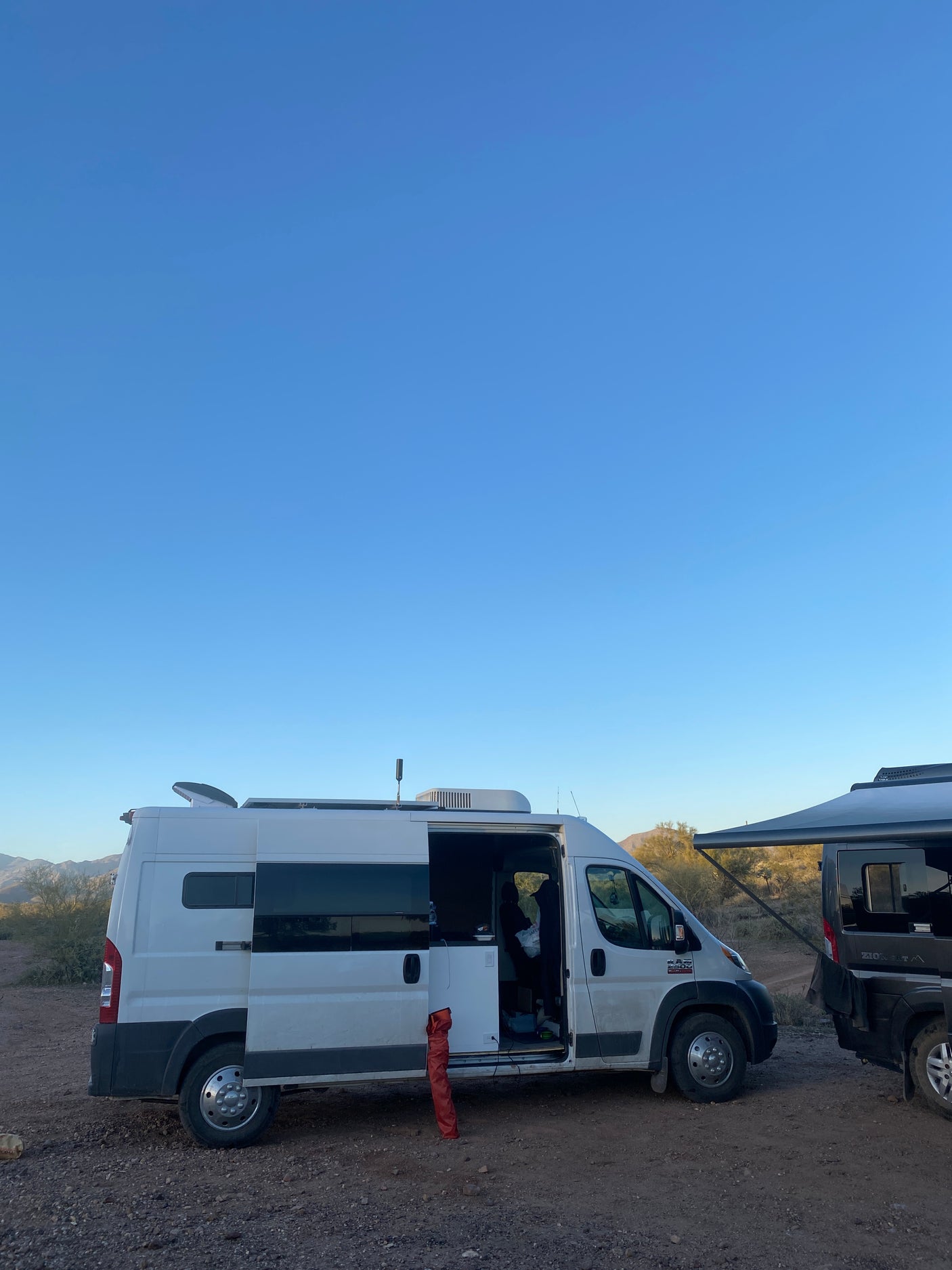White RV rental with side door open, parked next to another van in a sunny desert campsite. Ideal for your next outdoor adventure.
