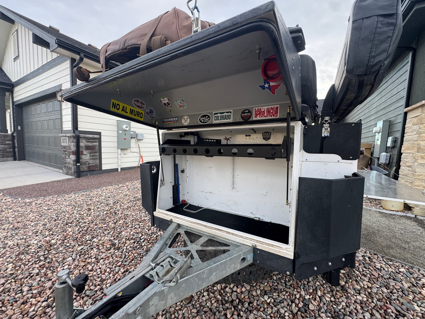 Open front storage on an off-road camper trailer, a type of RV rental, with various stickers on the lid. The trailer sits on a gravel driveway beside a house. — photo 16