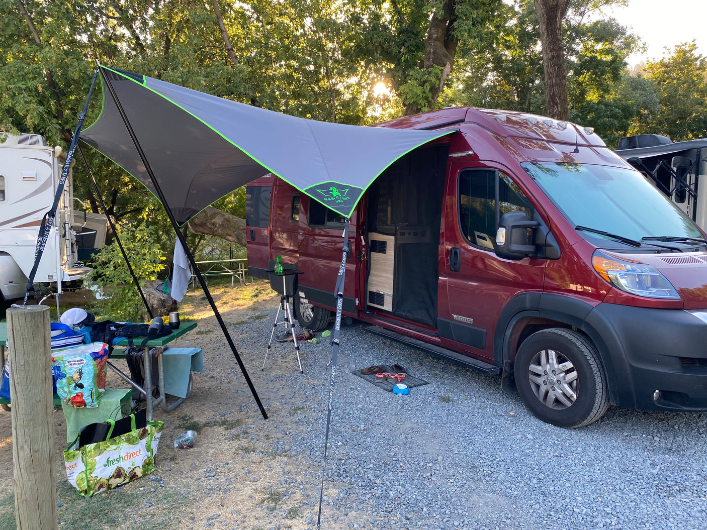 A red rv rental camper van with a large tarp awning set up at a shady campsite, ready for outdoor living.