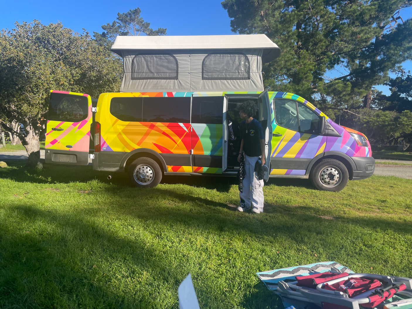 A colorful pop-top RV rental camper van on grass under a blue sky, with a person and camping gear visible.