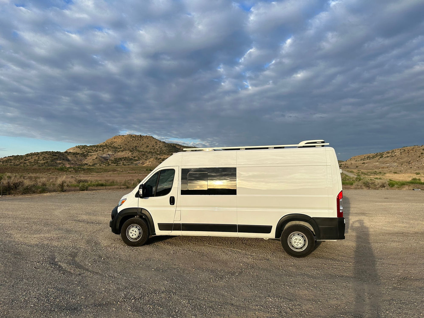 White RV rental van on a gravel lot, reflecting a vibrant sunset under a cloudy sky with hills.