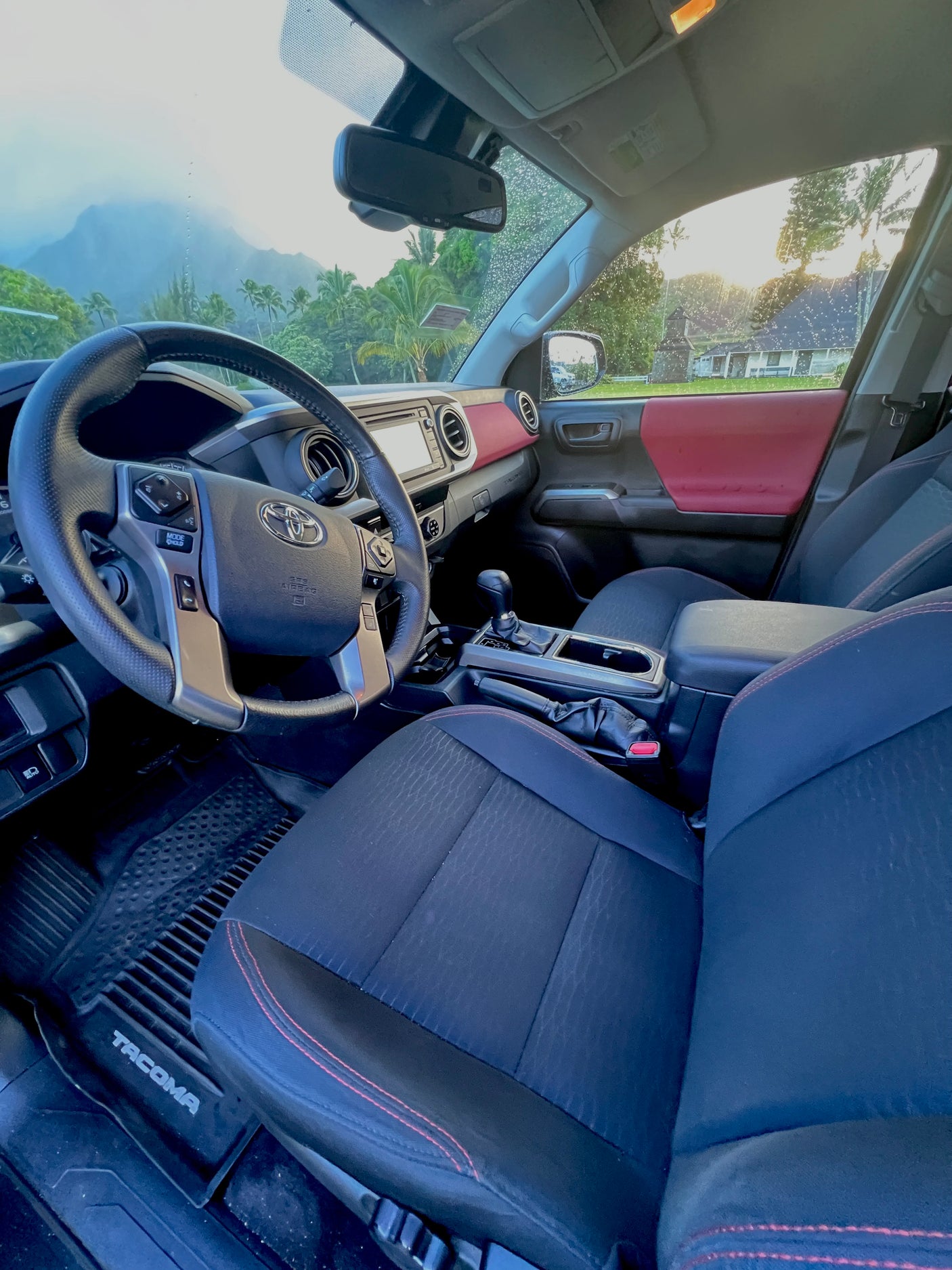 Driver's seat view of a vehicle interior with red accents. Tropical mountains and palms visible through the wet windshield. Great for an RV rental!