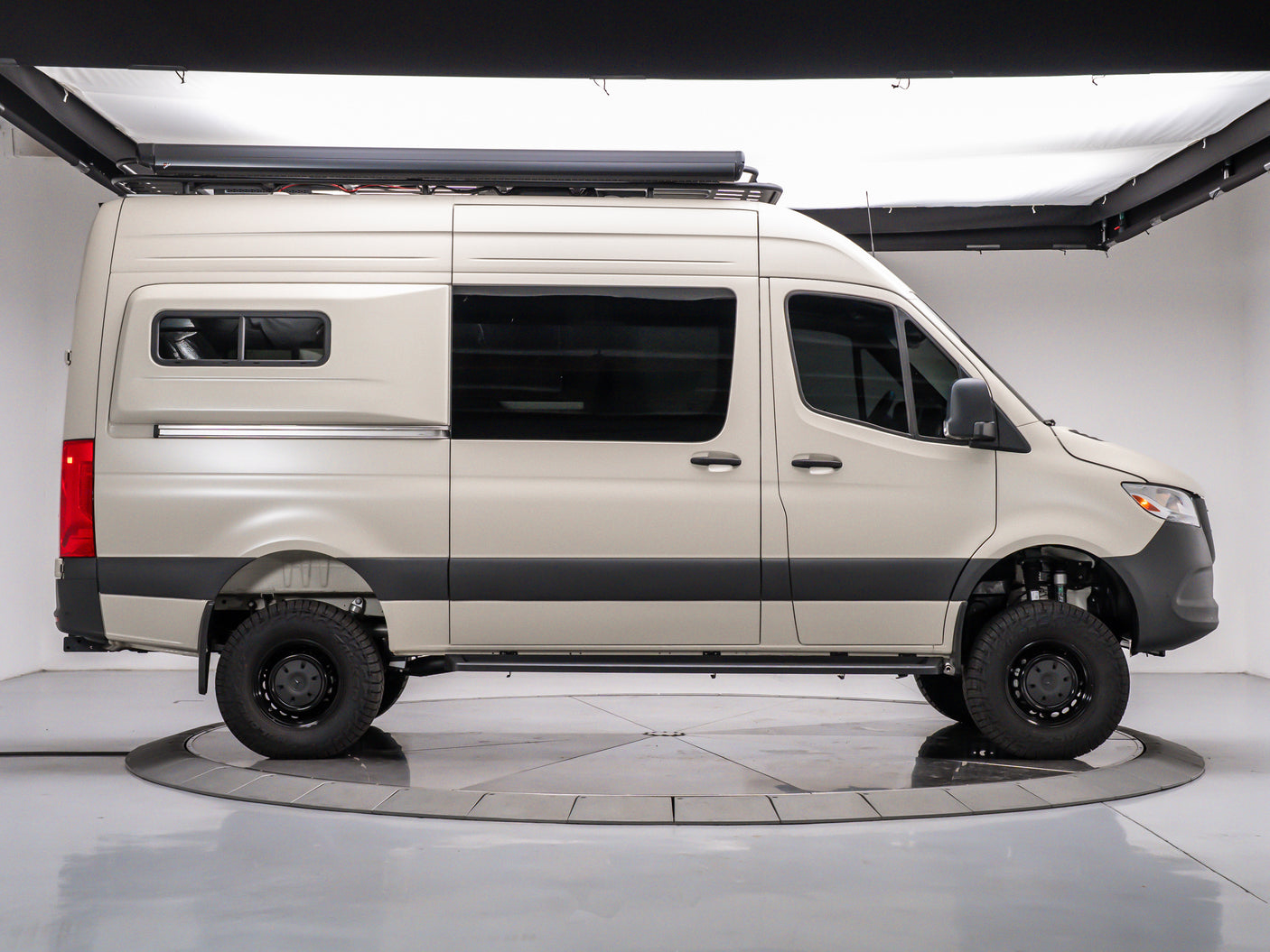 Side profile of a beige off-road camper van with a roof awning, chunky tires, and custom suspension, available for rv rental.