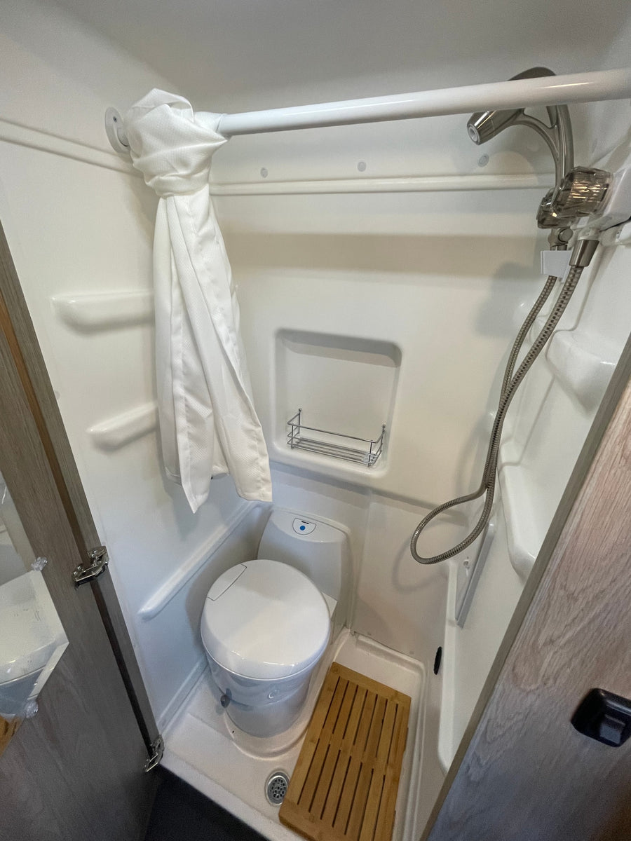 RV rental bathroom with a compact design, featuring a toilet, shower, curtain, shelf, and wooden floor mat.