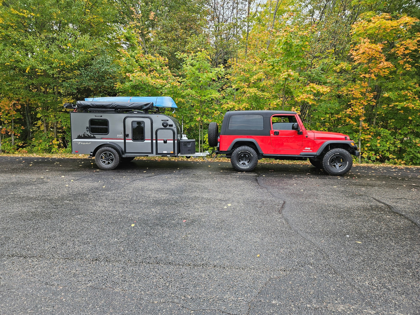 A red Jeep Wrangler tows a gray RV rental trailer with a blue canoe on top, parked in front of a forest displaying autumn colors.