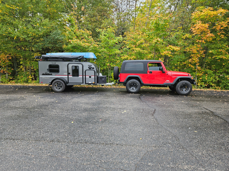 A red Jeep Wrangler tows a gray RV rental trailer with a blue canoe on top, parked in front of a forest displaying autumn colors.