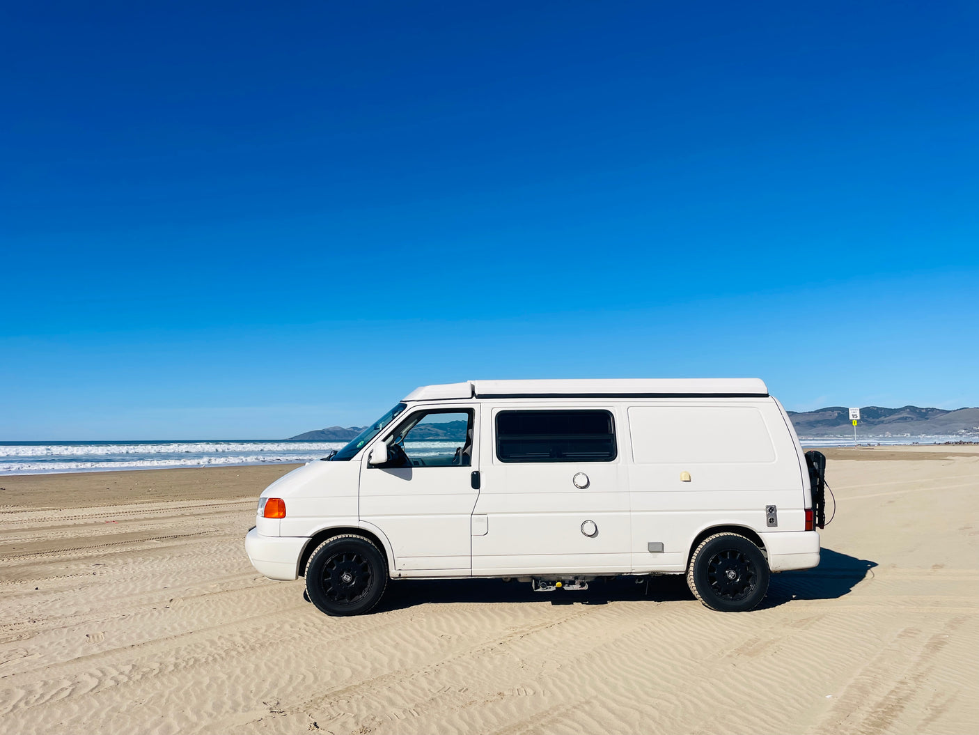 A white RV rental van sits on a sandy beach with ocean waves and clear blue sky, perfect for outdoor travel.