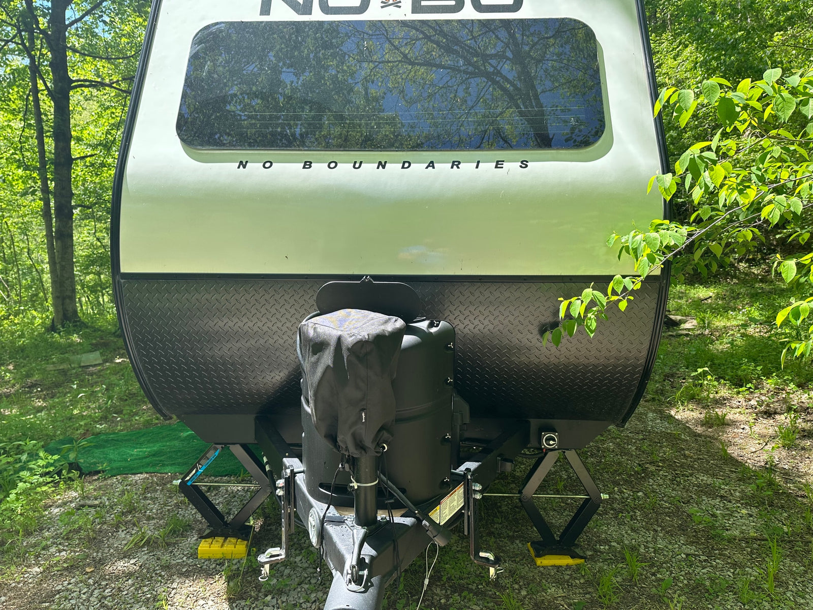 Front view of a "No Boundaries" travel trailer (RV rental) at a wooded campsite, showing tanks & deployed stabilizers.