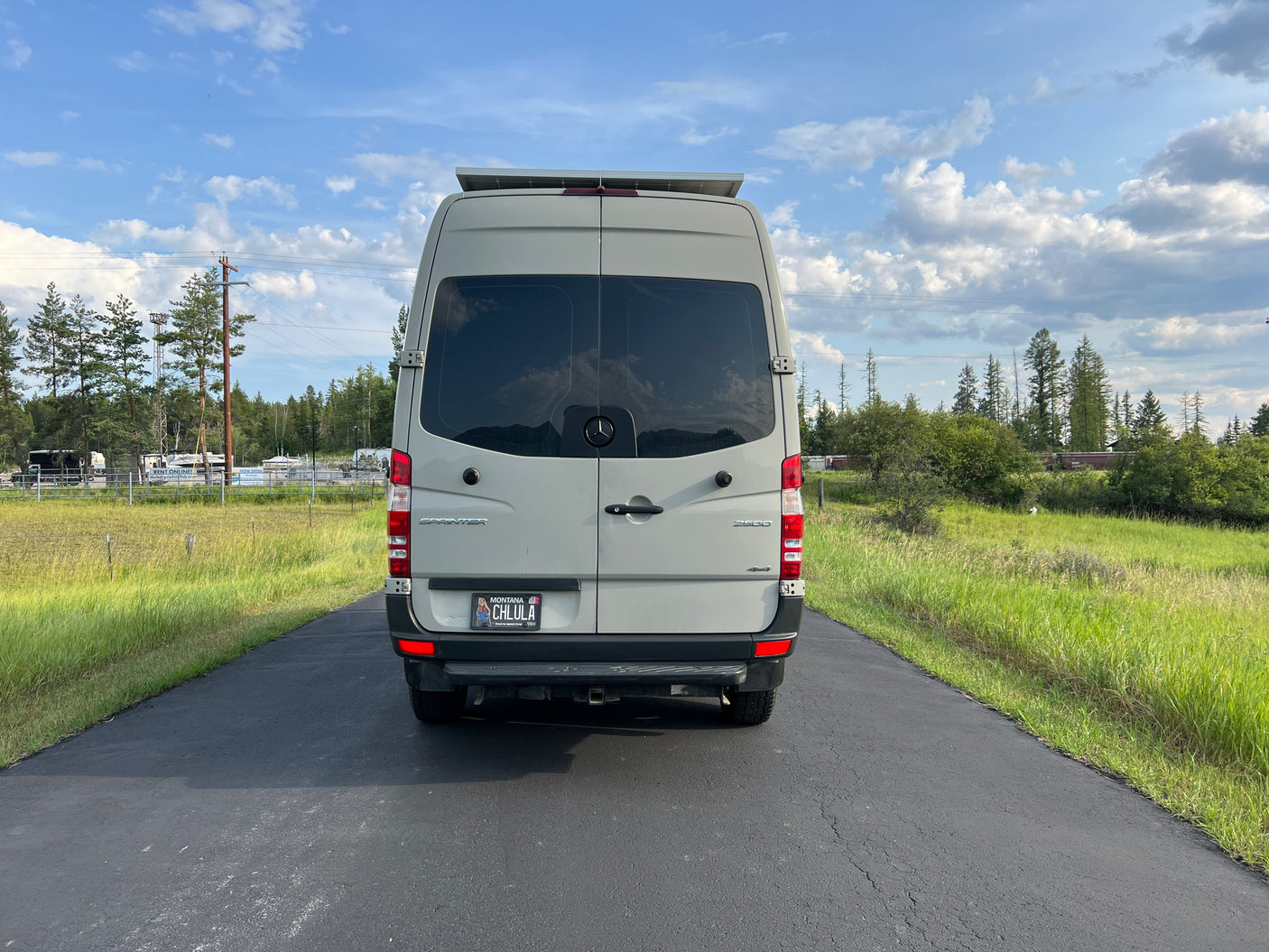 Rear of a grey Sprinter camper van, available for rv rental, on an asphalt driveway with trees and grass in the background.