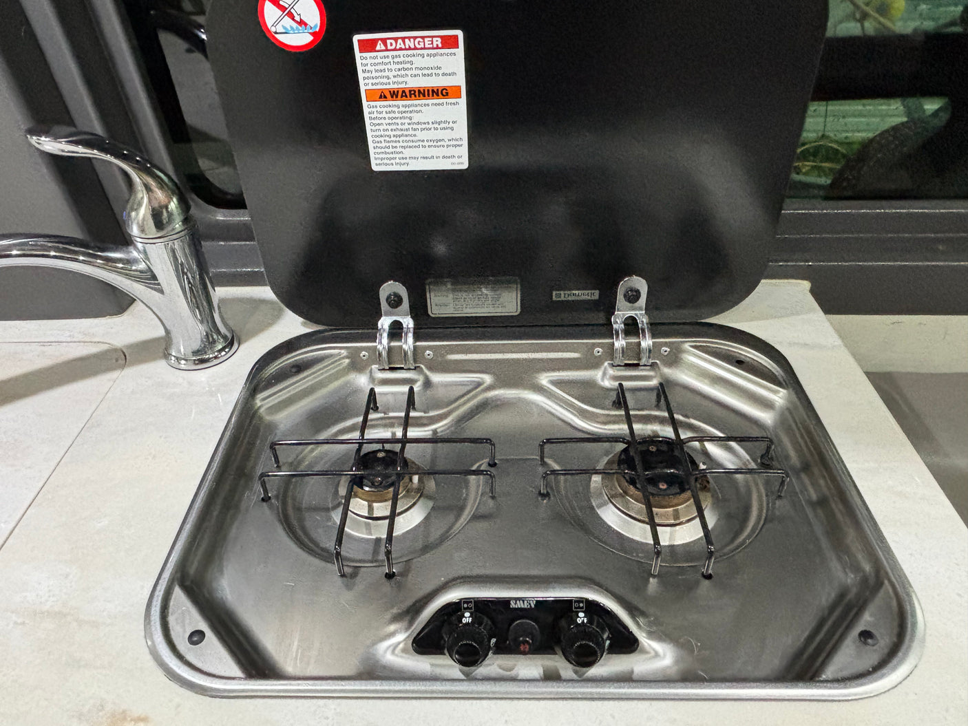 This RV rental kitchen features a compact two-burner gas stovetop with a safety warning on its cover and a chrome faucet.