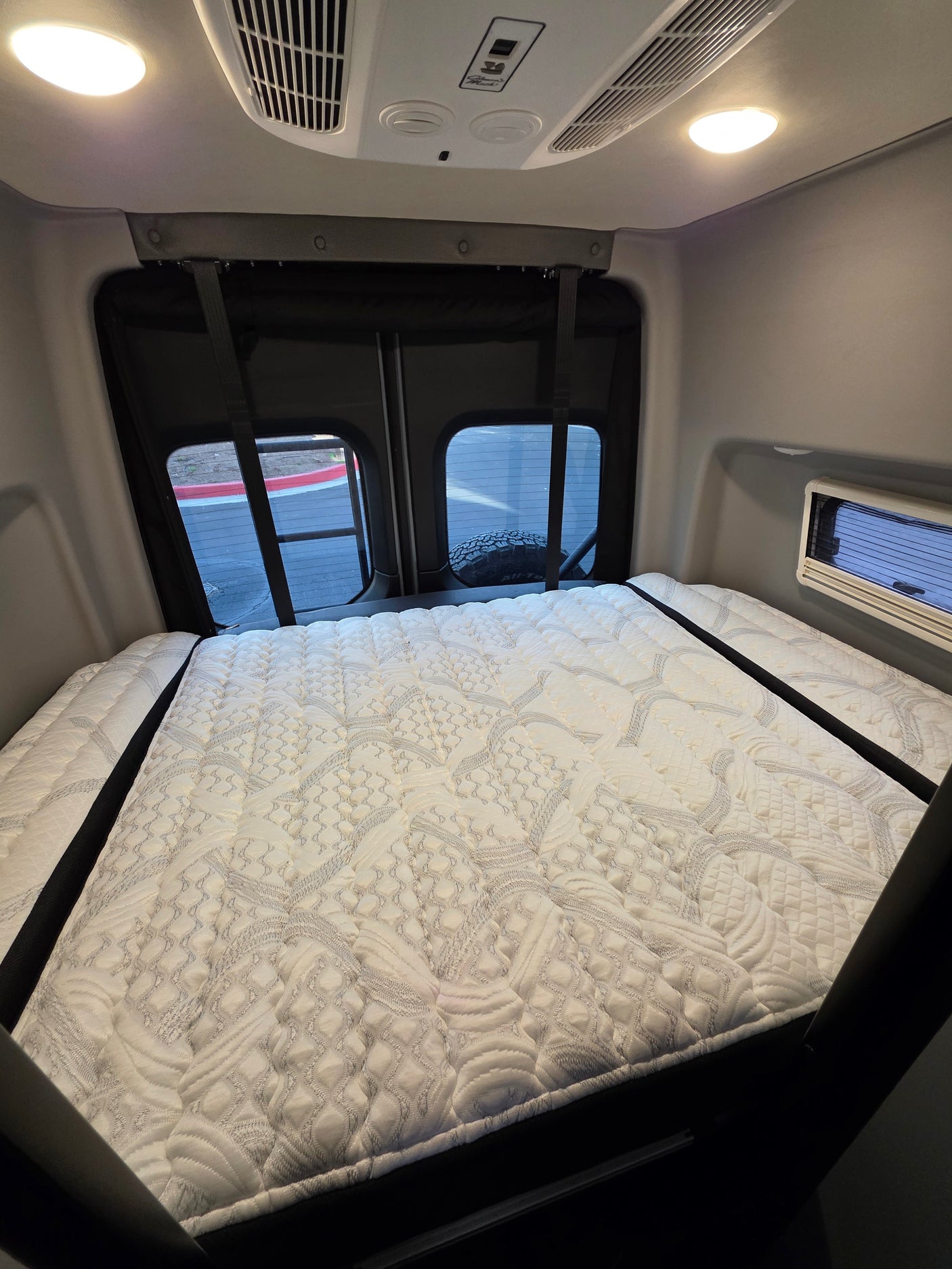Inside an RV rental: a plush, quilted mattress fills the rear sleeping area, flanked by windows. Ceiling features lights & AC.