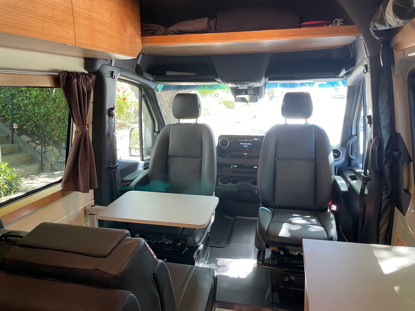 Bright RV rental interior showing swivel front seats, a table, and wooden overhead cabinets. Cozy camper for road trips.