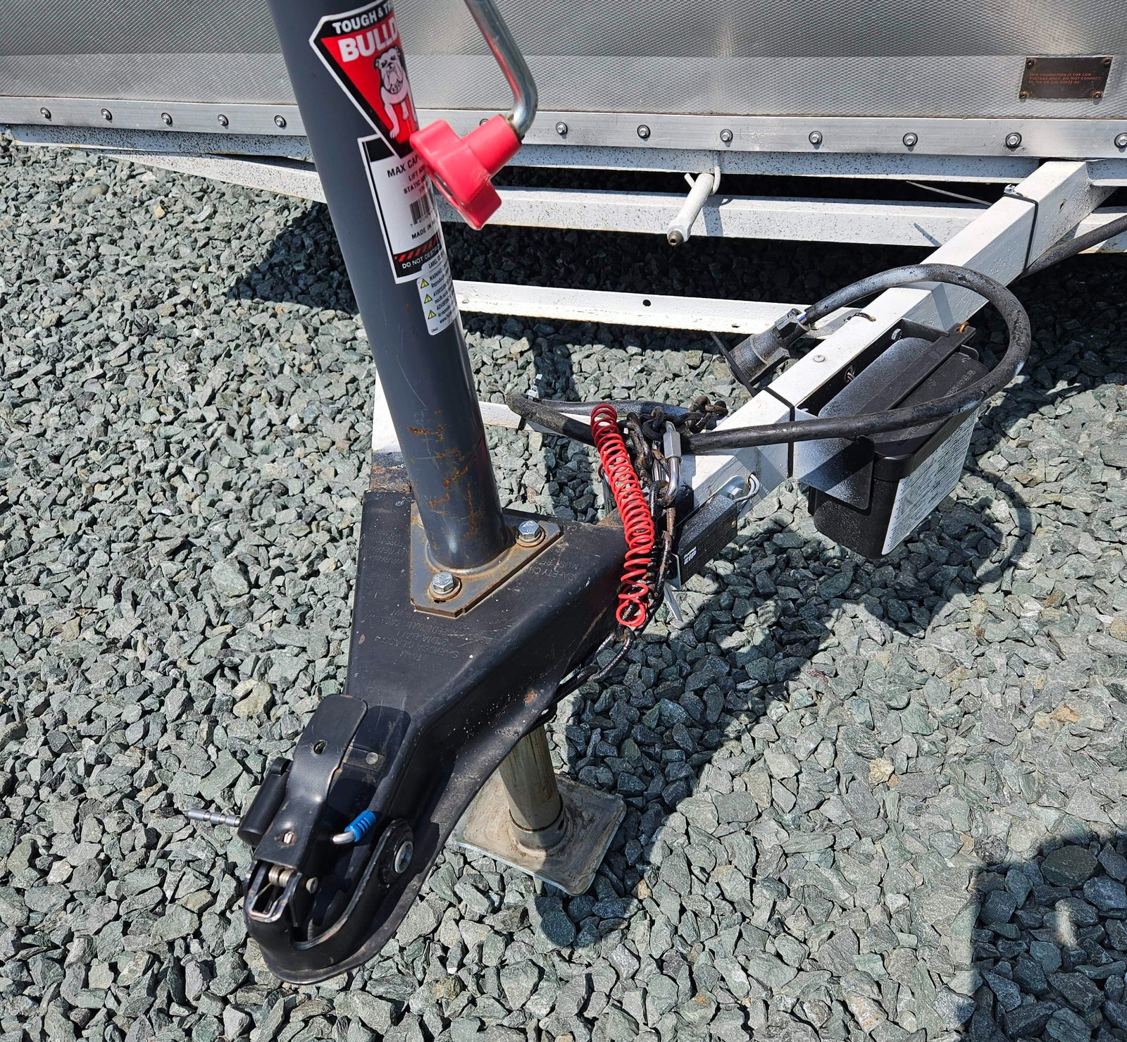 An RV rental trailer's hitch, jack, safety chains, and electrical wiring on gravel. A low voltage warning is visible.