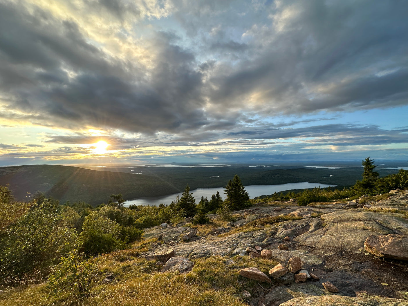 A vibrant sunset casts rays over a vast forested landscape and tranquil lake, ideal for an RV rental getaway.