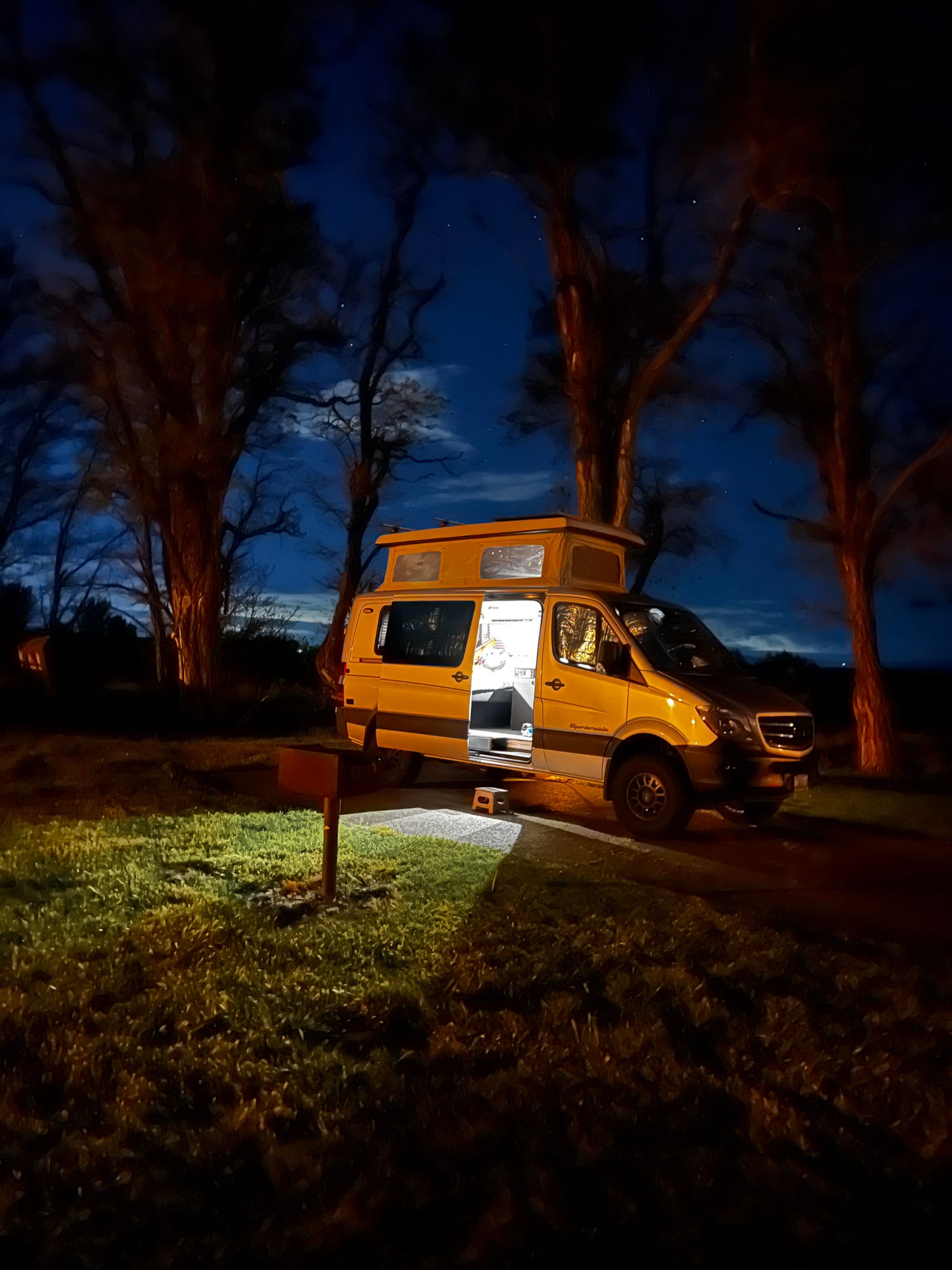 RV rental Sprinter camper van at a night campsite. Its interior glows, pop-top extended, under a starlit, tree-lined sky.